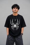 A man wearing a black short-sleeve t-shirt with a large white spider graphic on the front, paired with dark gray shorts, standing against a gray concrete wall.
