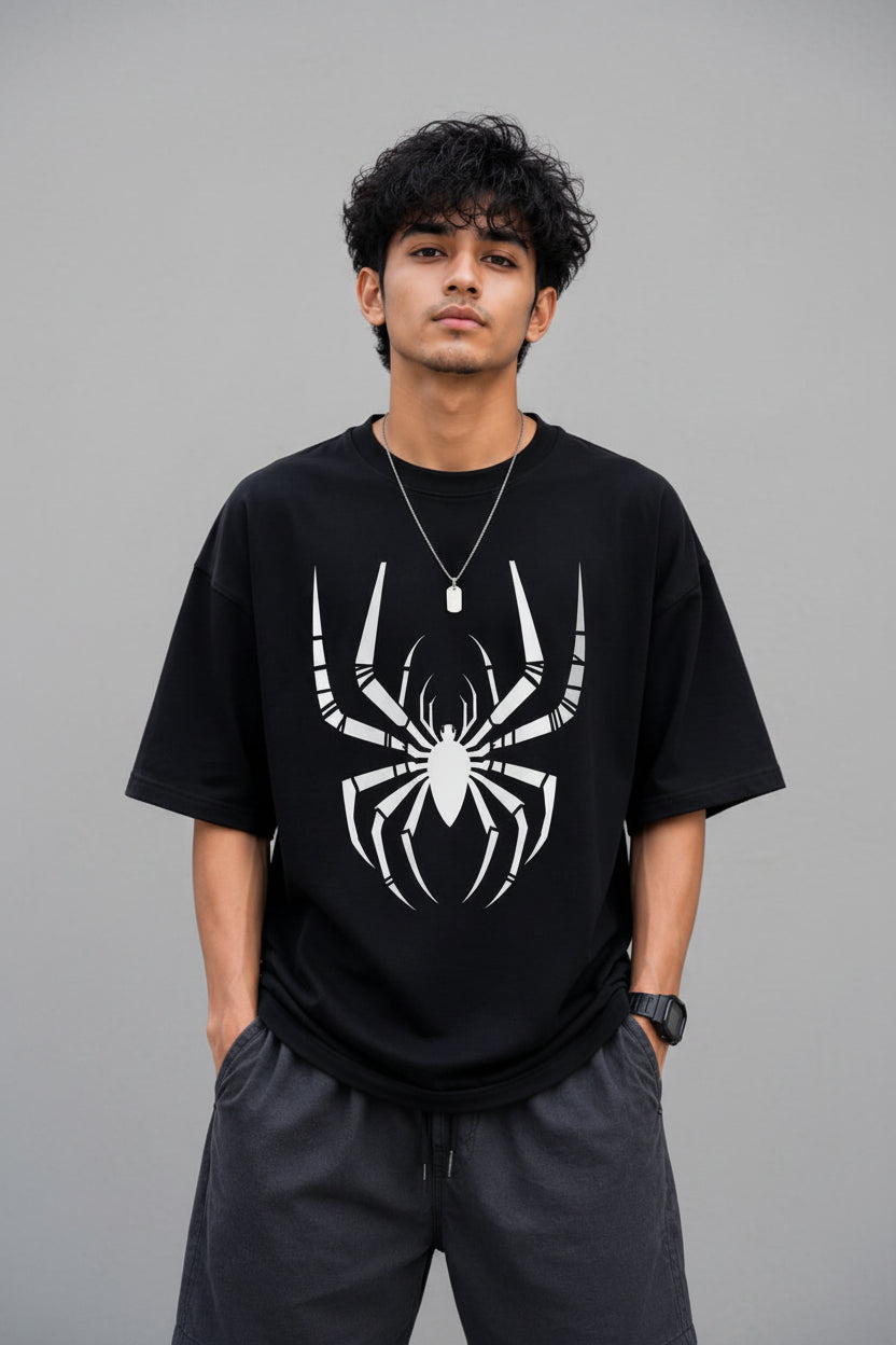 A man wearing a black short-sleeve t-shirt with a large white spider graphic on the front, paired with dark gray shorts, standing against a gray concrete wall.